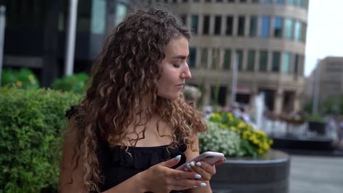 a Young Woman with Curly Hair Is Sitting on a City Street and Checking Her Phone