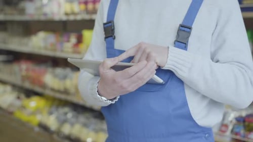 Unknown Caucasian Man in Uniform Standing in Grocery and Using Tablet. Unrecognizable Man Swiping