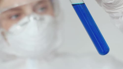A Lab Assistant Girl in a Medical Mask and Safety Glasses Holds a Flask of Blue Solution