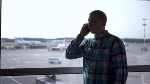A Young Man Is Talking on the Phone Against the Background of a Window at the Airport. Airplanes in