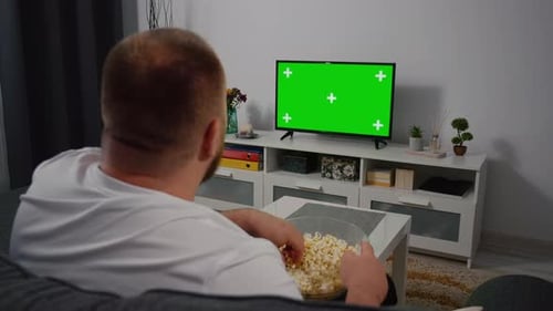 Man Watches Green Mock-up Screen TV while Sitting on a Couch at Home