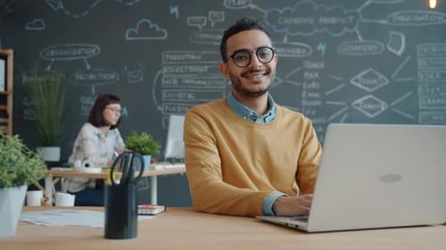 Porträt eines fröhlichen Mannes aus dem Nahen Osten am Schreibtisch mit Laptop im modernen Büro
