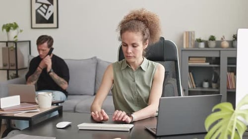 Woman Working at Computer Desk, Man on Sofa