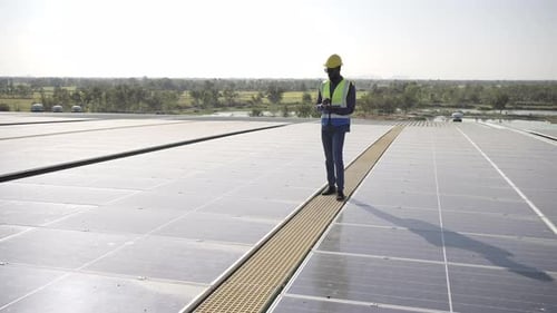 4K African man engineer using digital tablet maintaining solar cell panels on building rooftop.