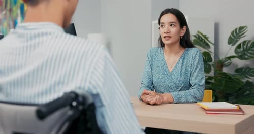A Job Interview with a Disabled Man in Wheelchair Beautiful Woman of Asian Korean Beauty Asks