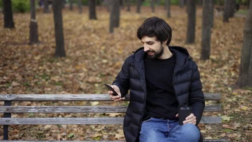 Brunette Man Sits on Bench in the Autumn Park Works on the Phone