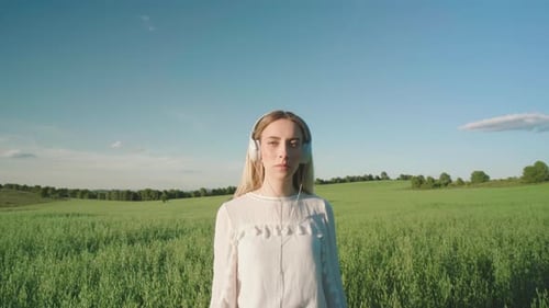 Pretty young woman with earphone listen music on green field during sunlight and blue sky. Portrait