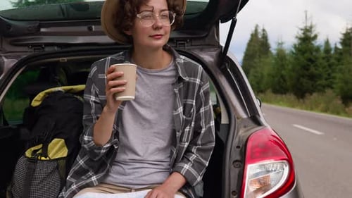 A Young Woman Drinks Coffee Sitting In The Trunk Of A Car. Girl Tourist Car Trip Woman Drinks Coffee