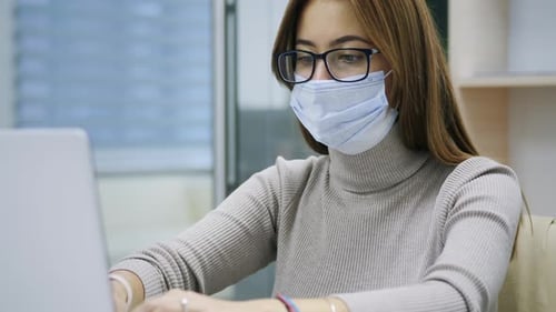 Female Employee in a Medical Facial Mask Works in the Office at the Computer. Work During