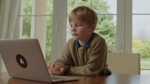 School Boy Using Laptop Sitting at the Table By the Window at Home