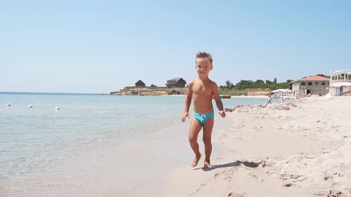 Boy with Light Hair Walks Along the Coast of the Purest Azure Sea on a Sunny Summer Day