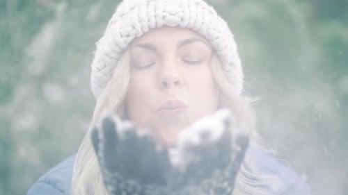 Happy Woman Blowing Snow From Palms with Mittens To Camera on Winter Forest Background