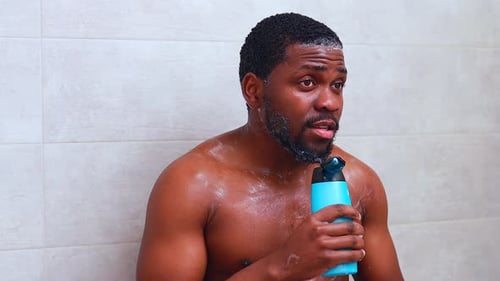 Attractive Young Cheerful Man Singing While Washing in the Shower Holding Shampoo Bottle