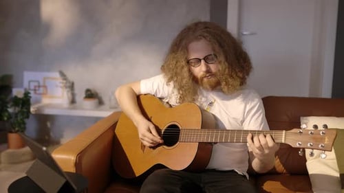 Joven aprendiendo a tocar la guitarra acústica viendo videos tutoriales en línea sentado en el sofá