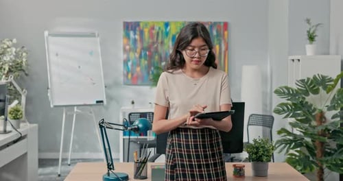 A Beautiful Woman with Glasses is Standing in Front of Desk in the Middle of the Office Checking on