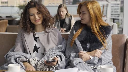 Two Caucasian Girlfriends Covered with Blankets Sitting in the Cafe Discussing Photos on