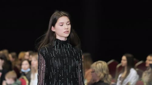 Brunette female model walk on podium. Girl in black dress with fringe pattern.