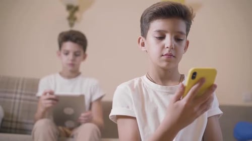 Close-up of Caucasian Boy Using Smart Phone As His Twin Brother Sitting at the Background