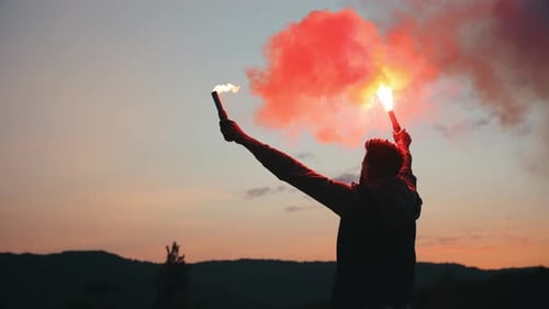 A Man is Standing on the Edge of a Cliff and Waving Two Flares in the Night