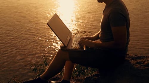 Young Adult Using Laptop by Water at Sunrise