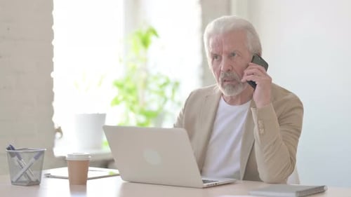 Alter Mann telefoniert, während er im Büro einen Laptop benutzt