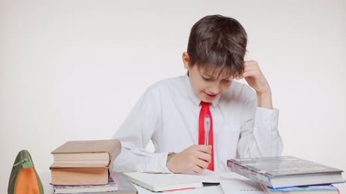 Thoughtful Caucasian School Kid in Red Tie Sitting at Table with Books Writing and Rejoicing on