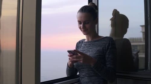 Woman Using Smartphone Near Window Overlooking Ocean