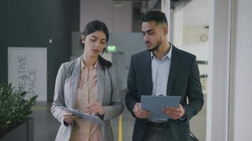Two Young Diverse Colleagues Walking Along Modern Office Building with Documents and Discussing