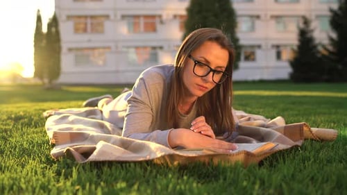 Niña con gafas leyendo libro acostada sobre una manta en el parque al atardecer
