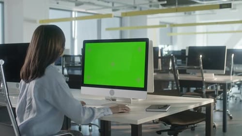 Asian Business Woman Typing on Desktop Computer with Green Mockup Screen While Sitting at His Desk