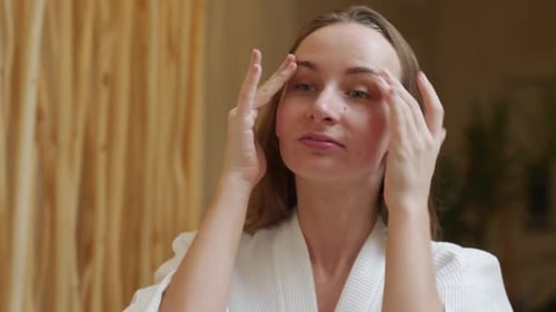 Lächelnde schöne Frau im Bademantel legt eine Hautpflegecreme auf ihr Gesicht und schaut ins Badezimmer