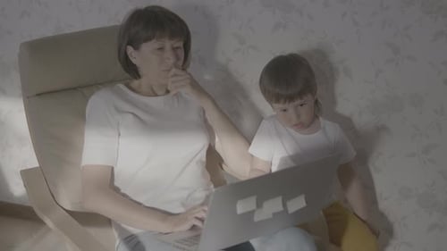 Mother and Her Toddler Boy Sit Together on Chair with Laptop. Woman Tries To Remote Work, but Kid Is