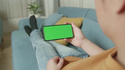 Male Using A Smartphone With Green Screen Mock Up Display At Home Living Room While Lying On Sofa