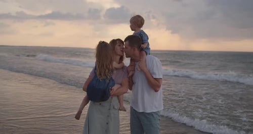 Young Parents Stand on the Sea Coast, Holding Their Little Children, Kissing