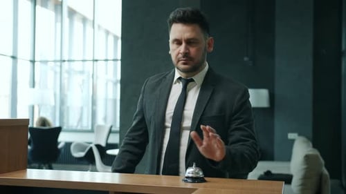 Middleaged Businessman Arriving at Hotel Ringing Bell at Reception Desk Talking To Manager