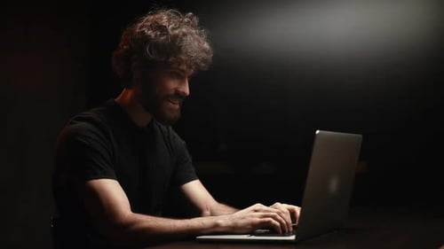 Side View of Smiling Young Man Typing Text on Laptop Sitting at the Desk in Dark Living Room