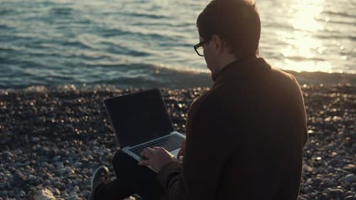 an Adult Man Sits on a Stone Beach Near the Sea and Picks Text on a Laptop