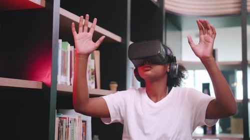 A schoolgirl sitting in front of library's bookshelf and using virtual reality glasses.