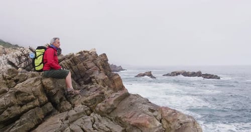 Senior hiker man with backpack sitting on the rocks while hiking near sea shore.