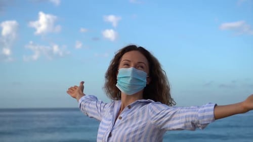 Glückliche Frau trägt medizinische Maske im Freien vor blauem Meer und Himmelshintergrund