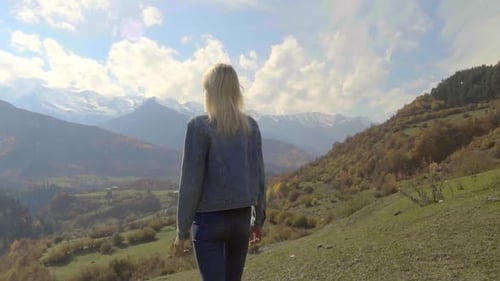 woman in denim jacket is standing on top of mountain, raising her hands in air,