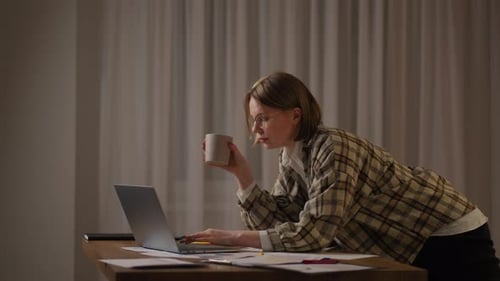 A Young Beautiful Business Woman Works From Home with a Cup of Coffee and Looks Into a Laptop Screen