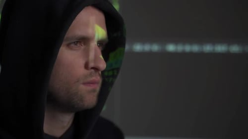 Male Hacker Working on a Computer While Green Code Characters Reflect on His Face in a Dark Office
