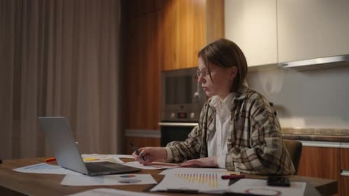 Side View A Woman with Glasses Works Remotely From Home Sitting at a Table with a Laptop and a