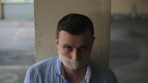 Businessman with His Mouth Sealed with Tape Sits Tied to Post Looks Up at Camera