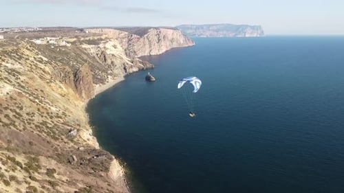 Aerial Drone View of a Man Flying a White and Blue Paraglider Over a Hill and Trees to the Sea Waves