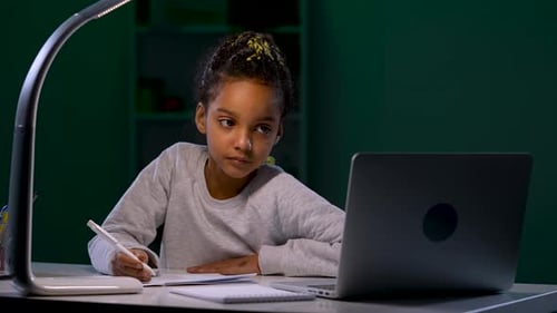 Una niña que usa una computadora portátil escucha un Vídeo tutorial y toma notas en su cuaderno
