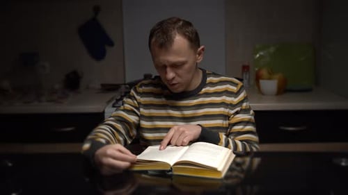 Man Reads Book at Table in Dim Room