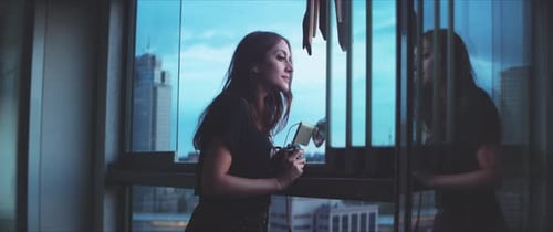 Woman Stands in City Apartment Looking Out Window