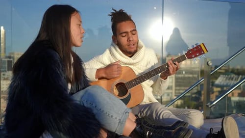 Porträt eines lächelnden Mannes, der Gitarre spielt, während Frau bei Sonnenuntergang auf dem Dach in der Stadt sitzt